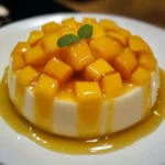 Gelatina de mango con queso crema servida en plato blanco, decorada con trozos de mango fresco.
